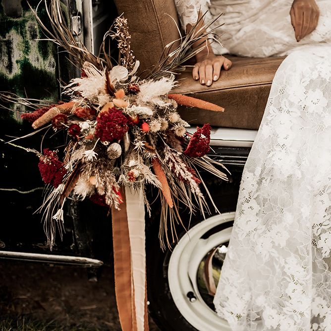 Bride in a lace wedding dress sitting in a VW Camper van with her red and dried flower bouquet 