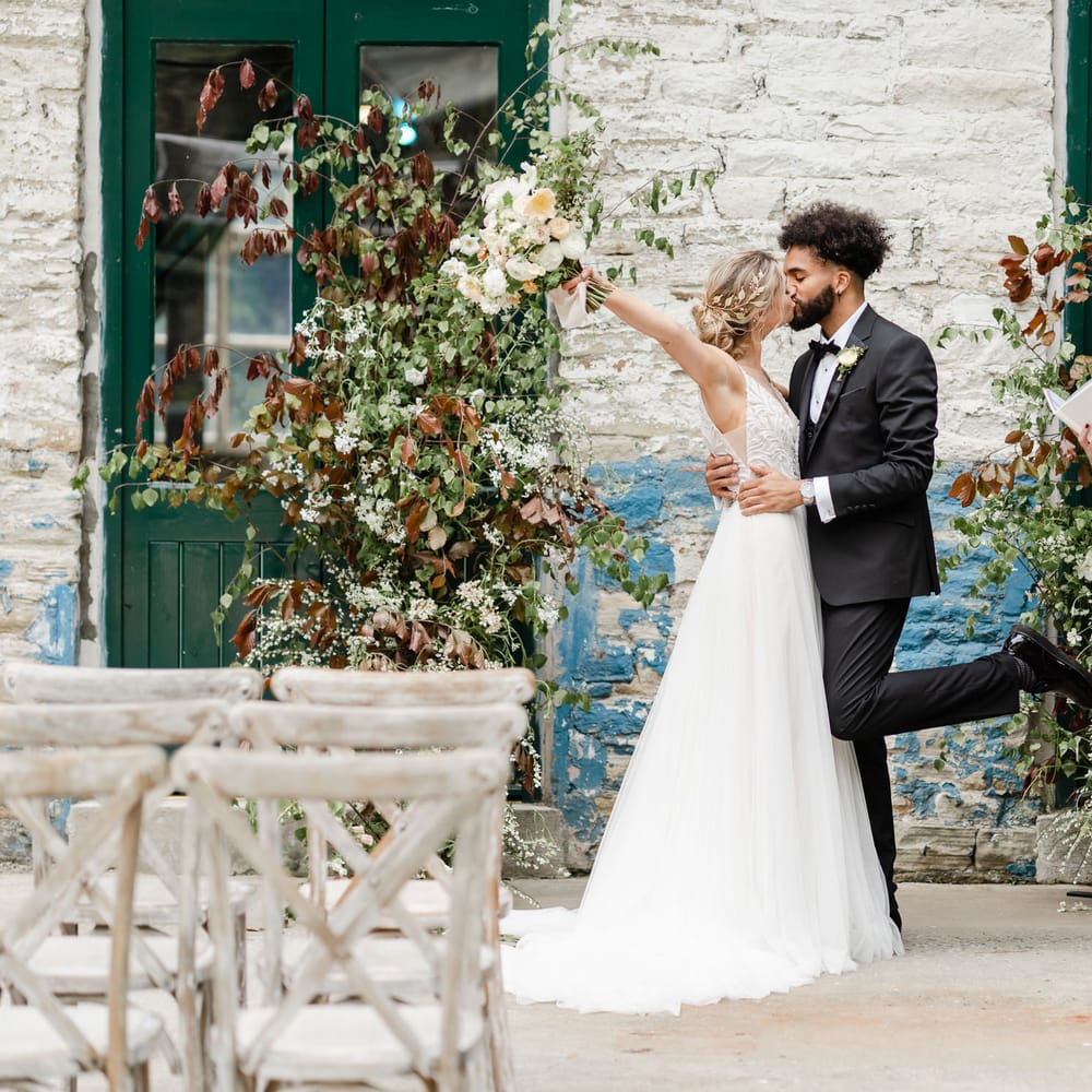 Groom in a tuxedo kissing his bride at the altar in a Maggie Sottero wedding dress as the celebrant watches smiling 