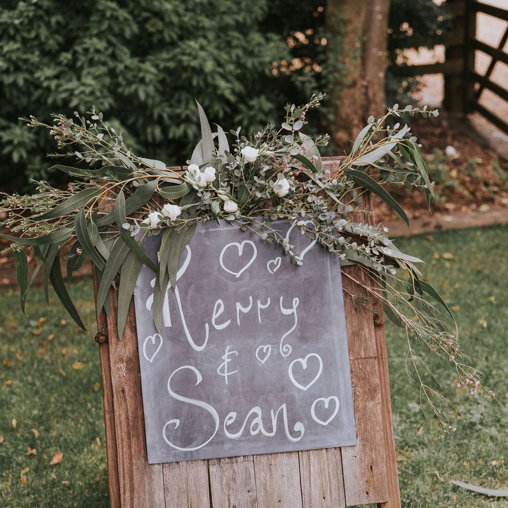 Wood and chalkboard wedding sign reading 'Kerry & Sean' topped with white florals and green foliage at Tythe Barn wedding with barn wedding flowers