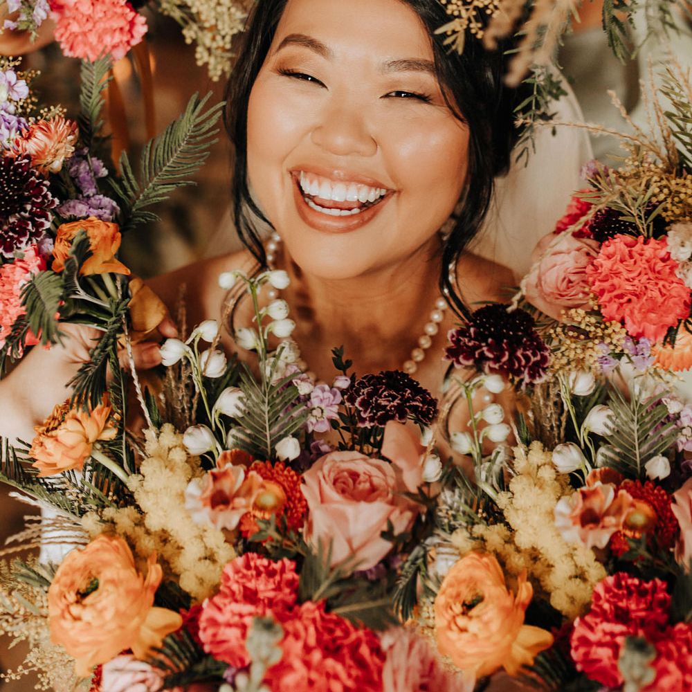 Bride with curled updo and pearl necklace smiles through hoop of colourful mixed florals for wedding at Scale in Liverpool