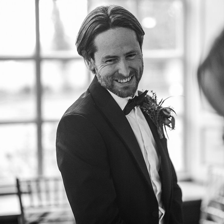 Groom wears black tie on his wedding day whilst smiling with wedding guests 