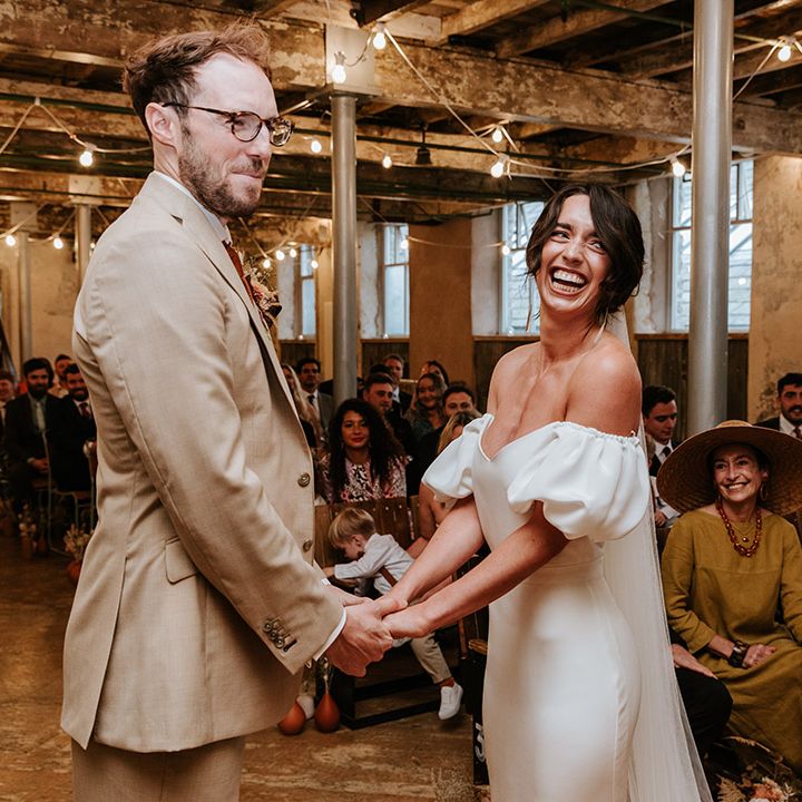 Groom in beige suit and brown glasses holds hands with the smiling bride in puff sleeve wedding dress at the altar