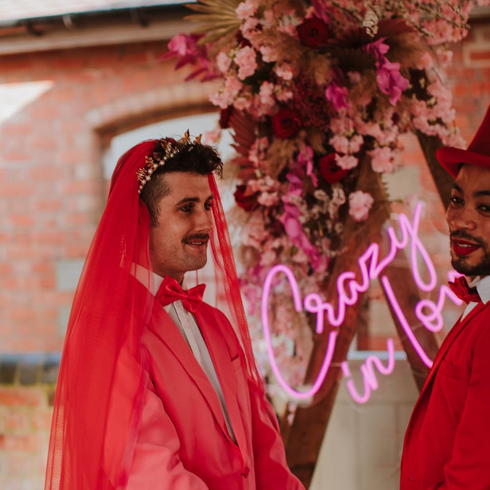 Stylish groom in a pink suit, gold crown and red veil holding hands with his groom at the altar 