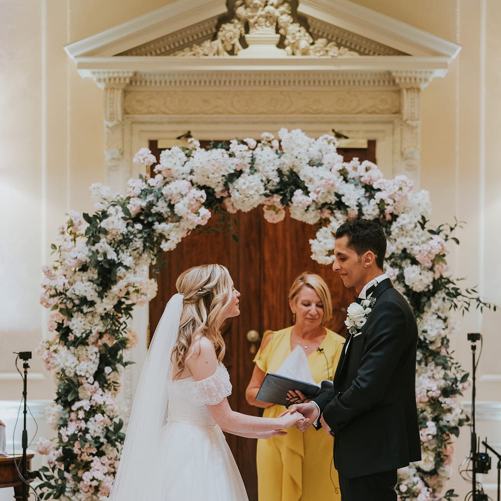 Bride and groom hold hands for their wedding ceremony in front of pink and white wedding flower arch 