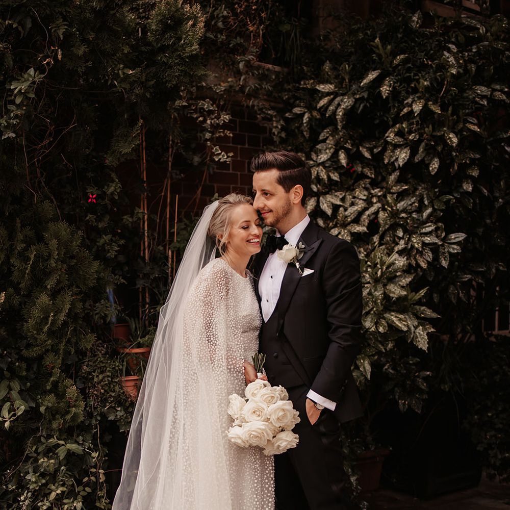 Bride in Jesus Peiro cape embraces the groom in black tie with white rose buttonhole and bouquet 