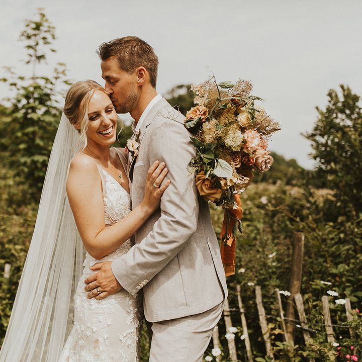Bride holding a pink and rust coloured wedding bouquet embracing the groom as he kisses her on the head 
