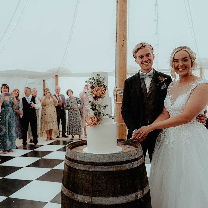 Three tier white iced wedding cake with orange and white flower decorations on a wooden barrel for marquee wedding reception 