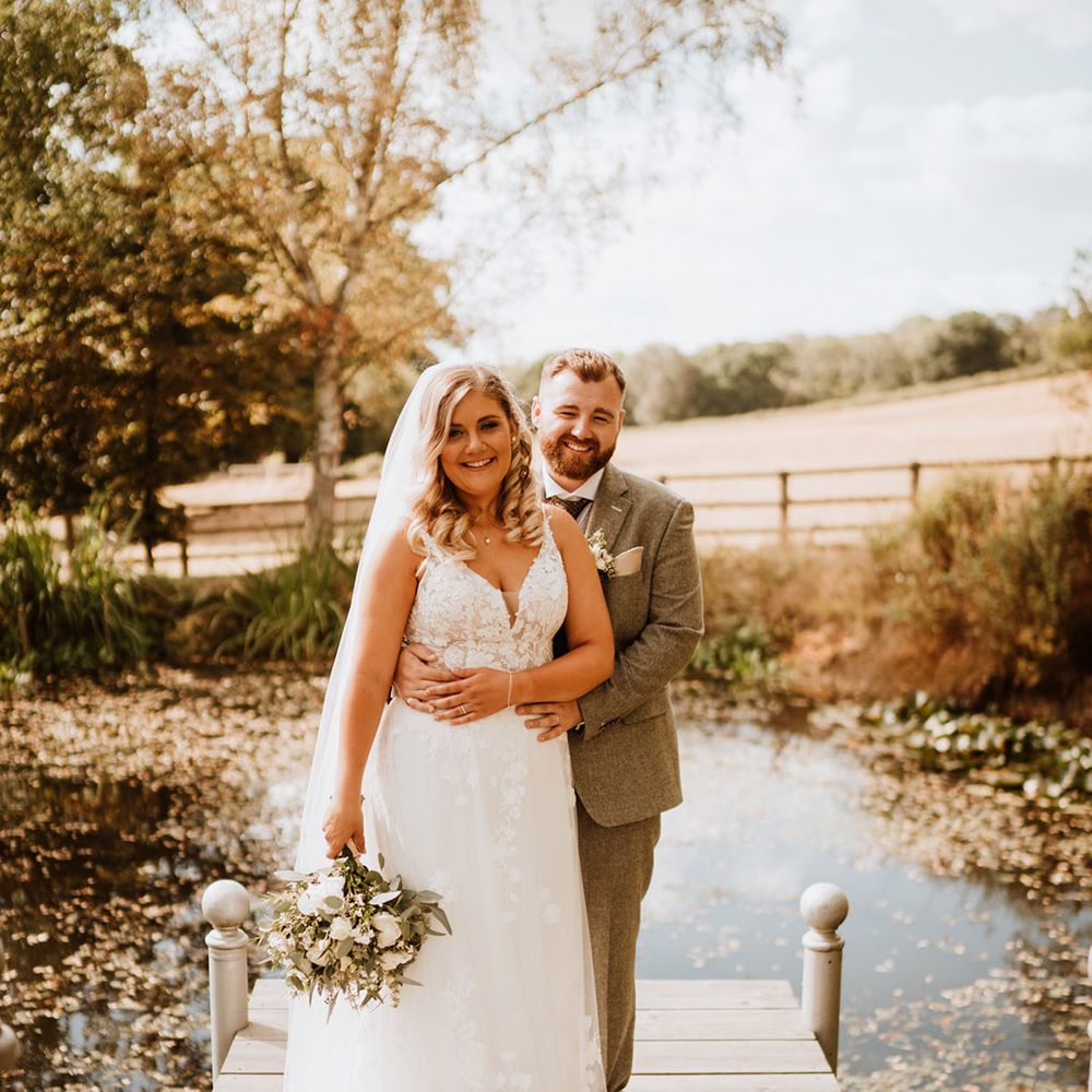The groom in a three piece tweed suit embraces the bride from behind at the stunning location surrounding The Oak Barn