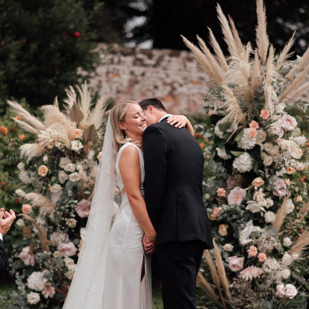 The groom kisses the bride on her neck as they stand at a the altar for their ceremony in front of large flower columns with pampas grass 