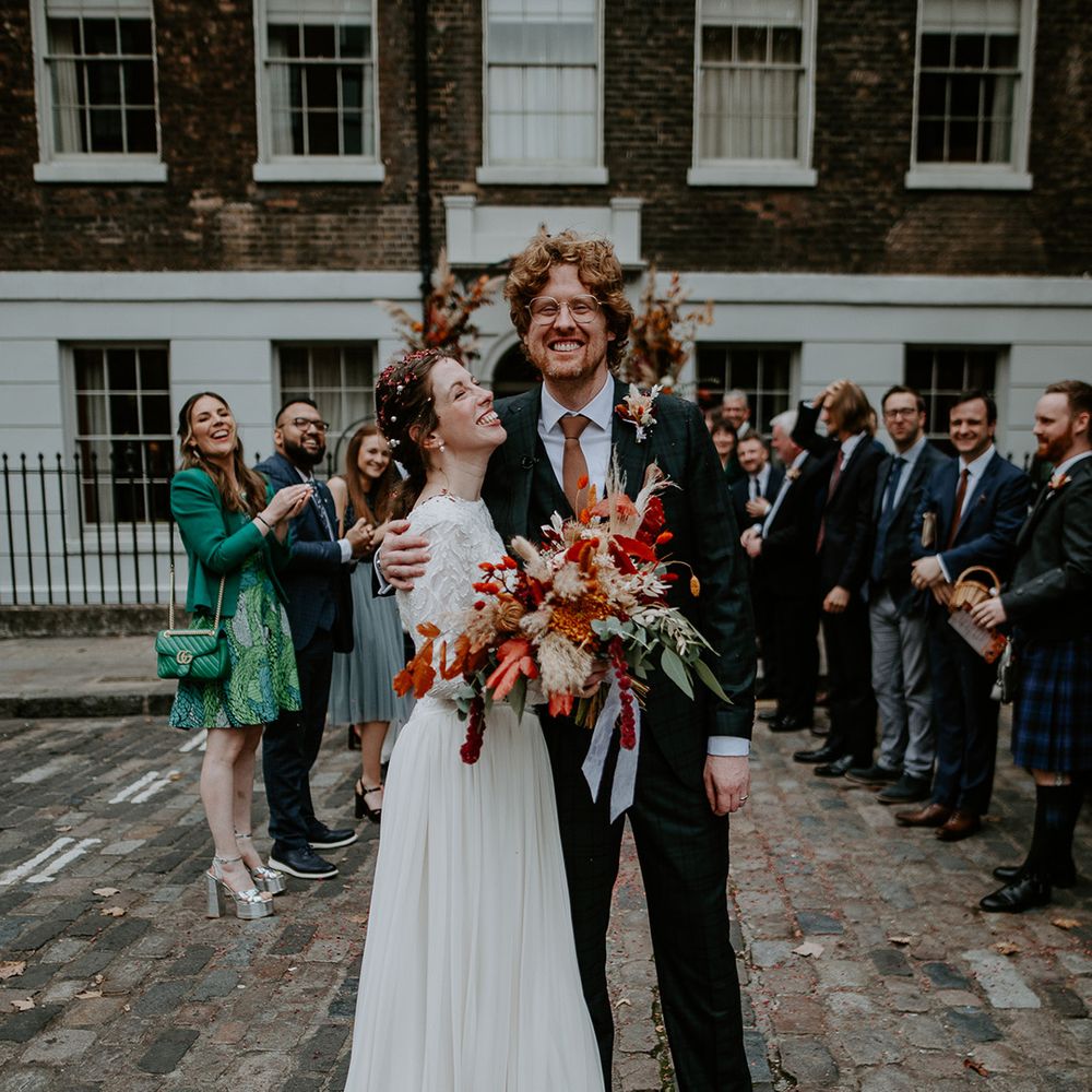 Groom in dark suit with orange tie and autumnal boutonniere embracing bride in long sleeve lace vintage wedding dress and closed toe satin heels holding large autumnal bouquet with orange, black, green and yellow roses, peonies, foliage and pampas grass surrounded by wedding guests on the street