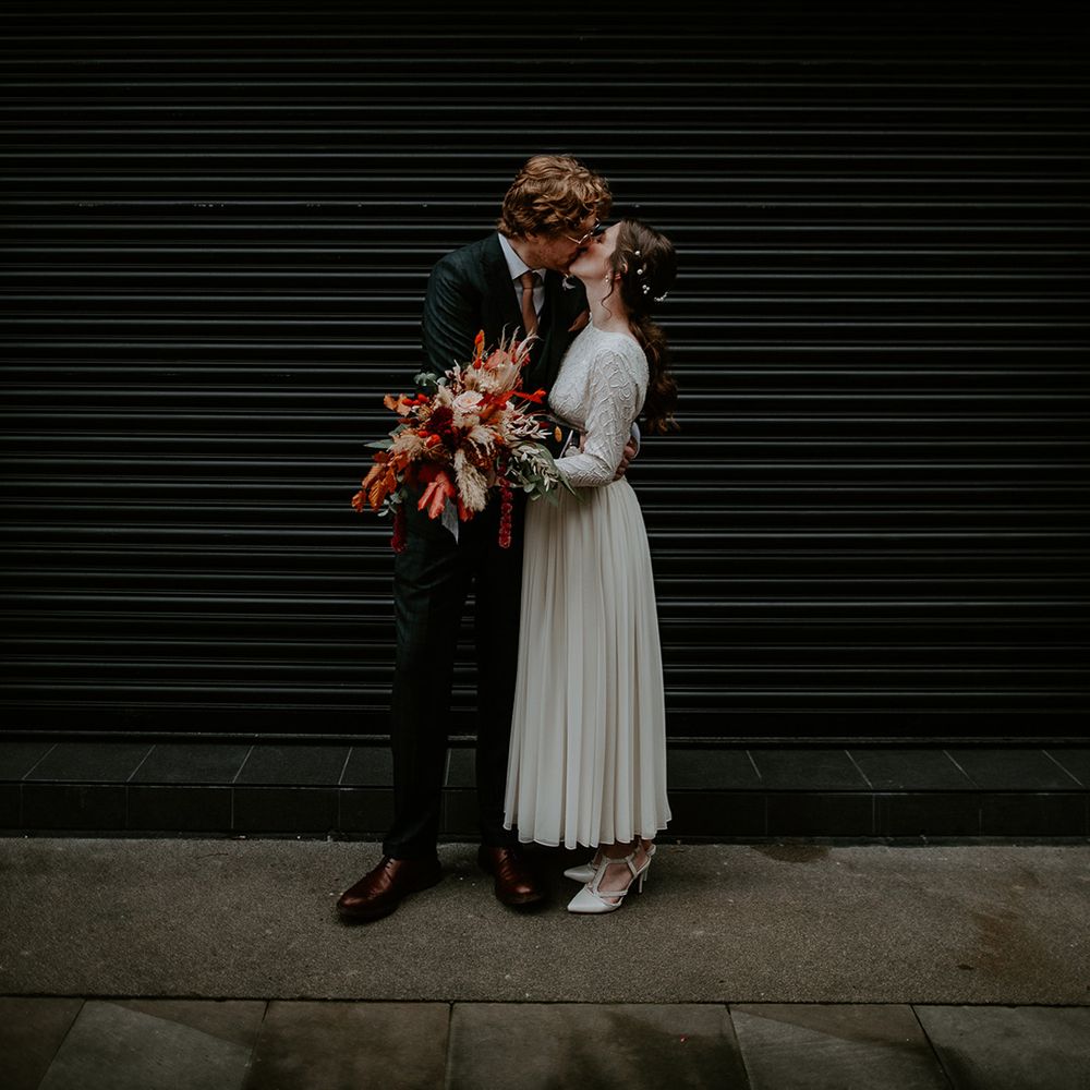 Groom in dark suit with orange tie and autumnal boutonniere kissing bride in long sleeve lace vintage wedding dress and closed toe satin heels holding large autumnal bouquet with orange, black, green and yellow roses, peonies, foliage and pampas grass