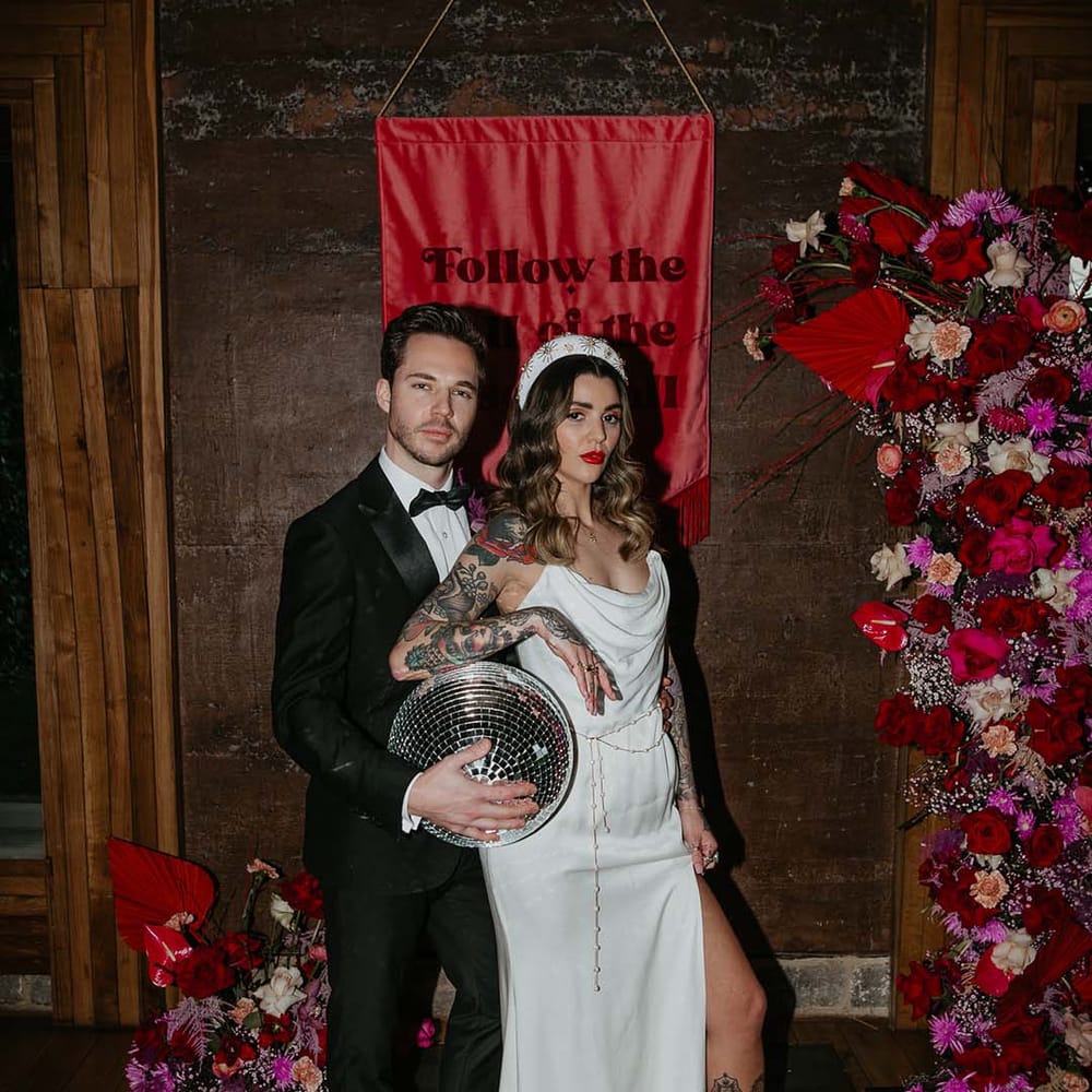 Groom in classic black tuxedo with magenta amaryllis and orchid boutonniere and bride in cowl neck sleeveless satin wedding dress with leg slit, gold glittery wedding headband posing with large wedding disco ball decor