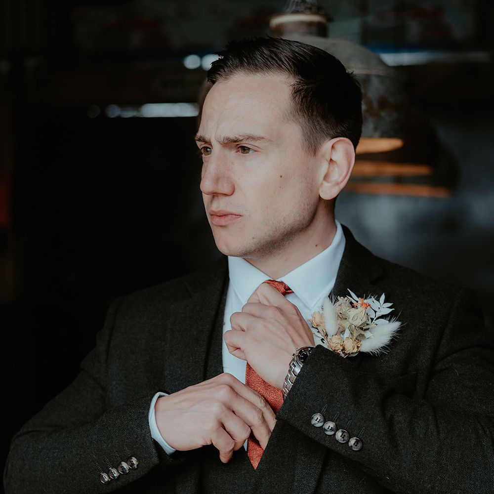 The groom in a black suit with an orange tie wearing a dried flower and bunny grass buttonhole for the rustic glam wedding 