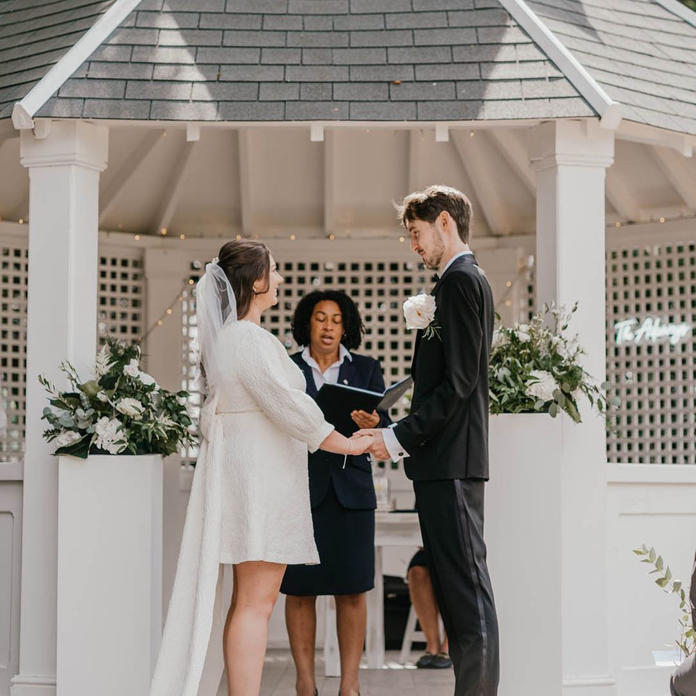 The groom in traditional black tuxedo stands holding hands facing the brides in a short wedding dress for their outdoor gazebo wedding 