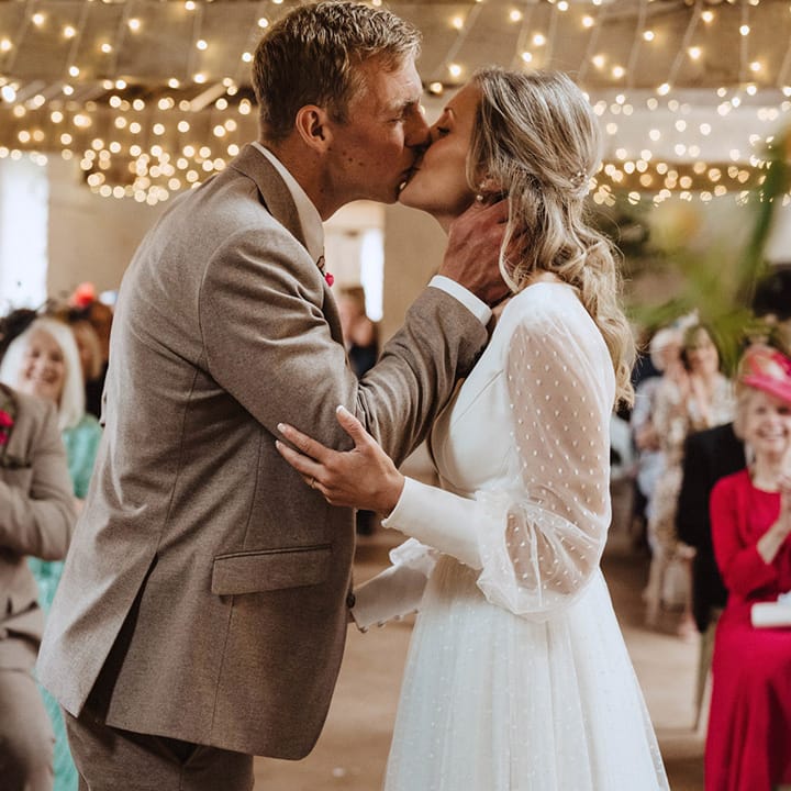 The groom in a beige three piece suit and the bride in a long polka dot sleeve Sassi Holford wedding dress share their first kiss as a married couple