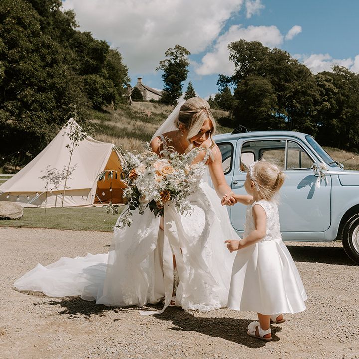 The bride greets a flower girl in a white dress with blonde hair in a ponytail next to the baby blue wedding car Fiat 500 