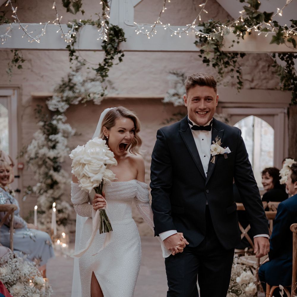 Groom in traditional black tie with the bride in sparkly off the shoulder wedding dress holding white rose bouquet of flowers 