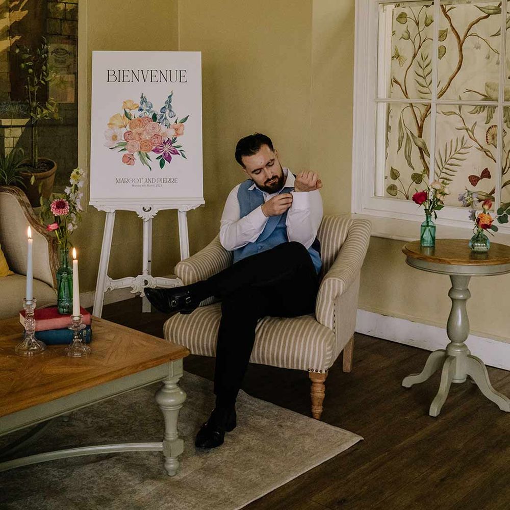 Groom wearing cornflower blue waistcoat and black suit trousers fixing his shirt in the reception room of Braxted Park wedding venue 