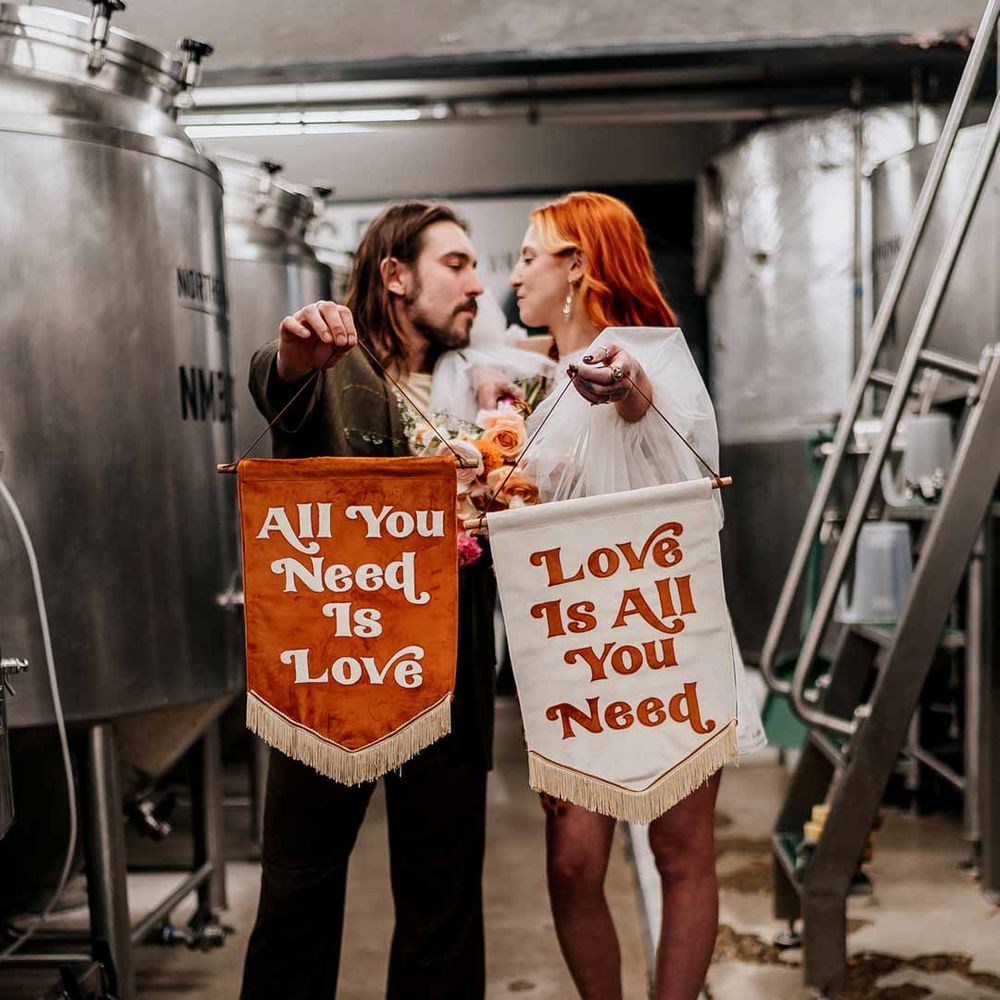 Bride and groom holding up velvet fabric wedding banners with retro lettering in burnt orange and white standing by beer fermenters at Northern Monk Refectory Leeds