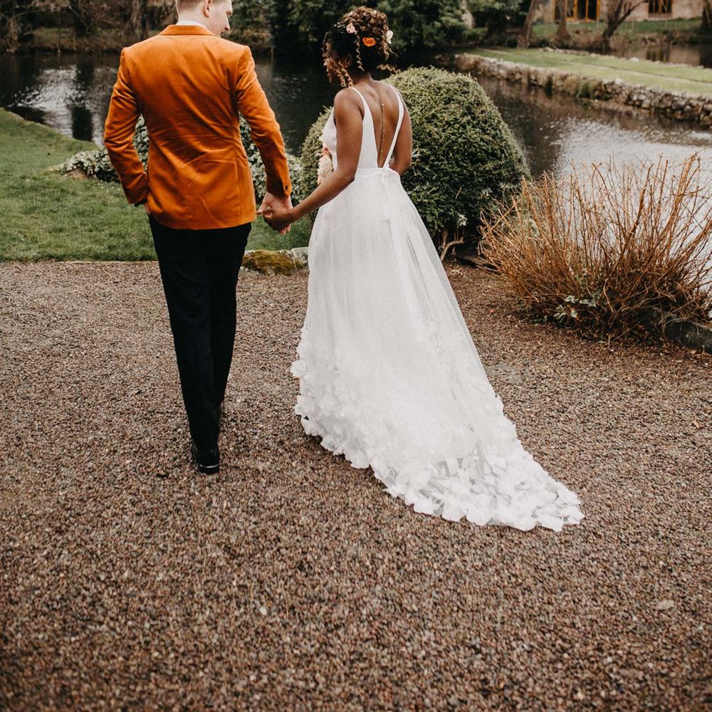 Bride in low back sleeveless wedding dress with 3D floral applique train walking with groom in crushed burnt orange velvet grooms blazer