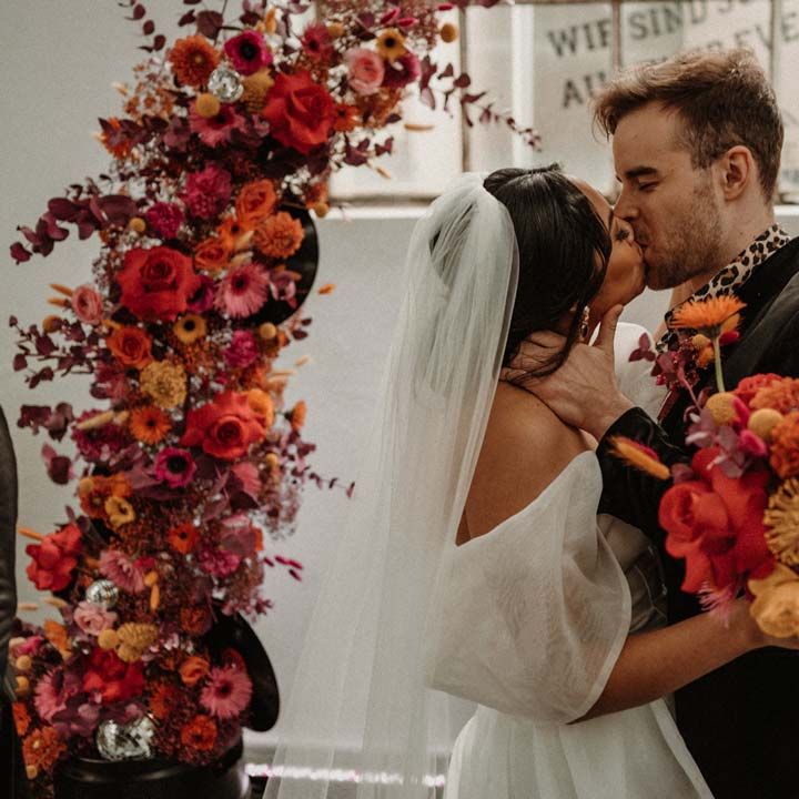 Bride in off shoulder tulle short wedding dress with church length veil kissing groom wearing wet look black grooms suit with leopard print shirt and pink and orange boutonniere under vinyl wedding arch