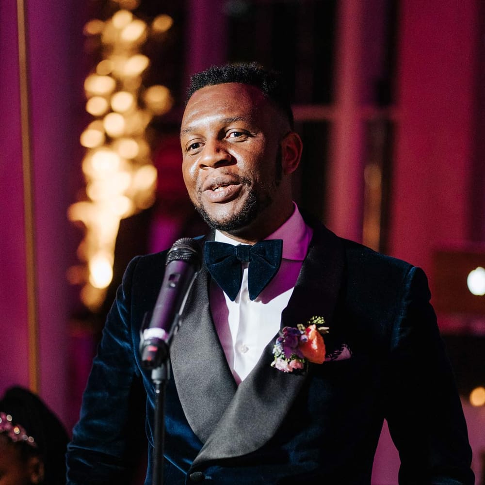 Groom in velvet midnight blue double breasted groom’s blazer, velvet bowtie and peach garden rose boutonniere doing grooms speech for vow renewal ceremony at Syon Park 
