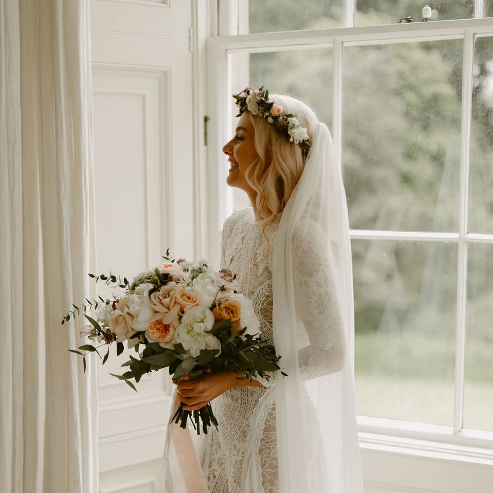 Bride in lace wedding dress holding wedding bouquet standing in front of window with white curtains