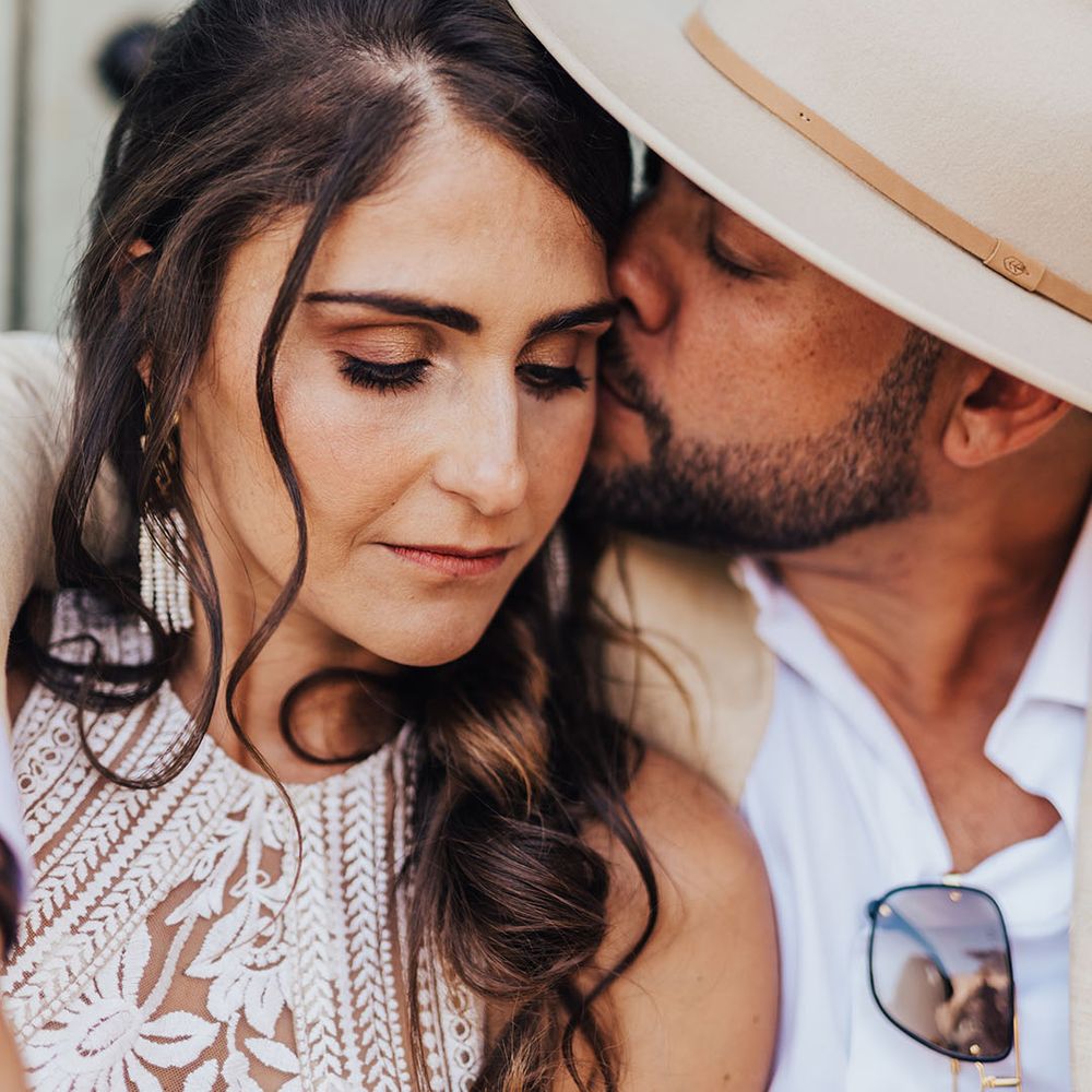 Bride with ponytail hairstyle being kissed on the cheek by the groom in a hat 