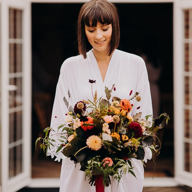 Bride with bob and fringe on the wedding morning in a white getting ready robe holding her colourful bouquet 