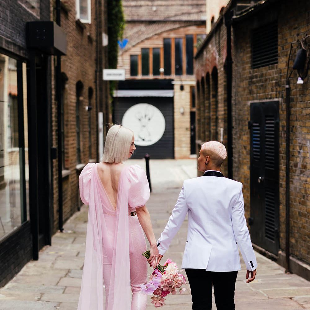 Bride in a custom-made pink jumpsuit with slim leg trousers and puff sleeves holding hands with a her bride in a white tuxedo jacket 