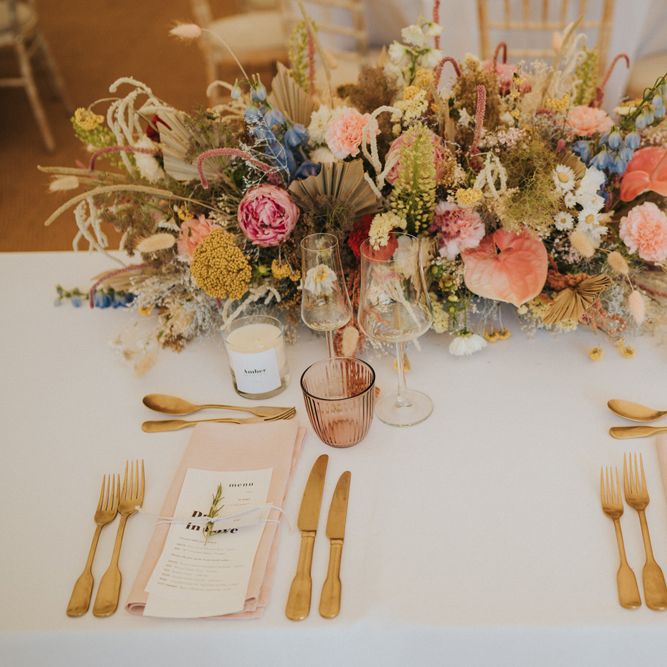 Marquee reception place setting with gold cutlery, pink napkins, and a top table flower arrangement with dried palm leaves and anthuriums 