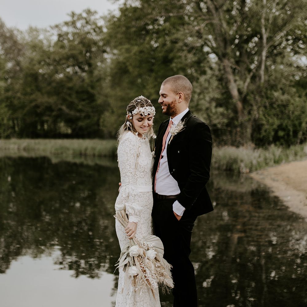 Bride in lace wedding dress holding a dried palm leaf bouquet standing next to the lake at Waresley Park Estate 