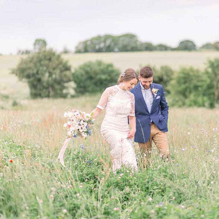 Bride in a lace Phase Eight wedding dress walking though the fields with here groom in a navy blazer 