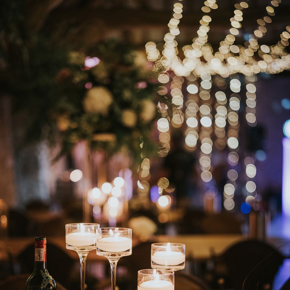 Glass long stem candle holders next to flowers in small gold vases and bottle red wine in fairy lit room at Tythe Barn wedding with barn wedding flowers