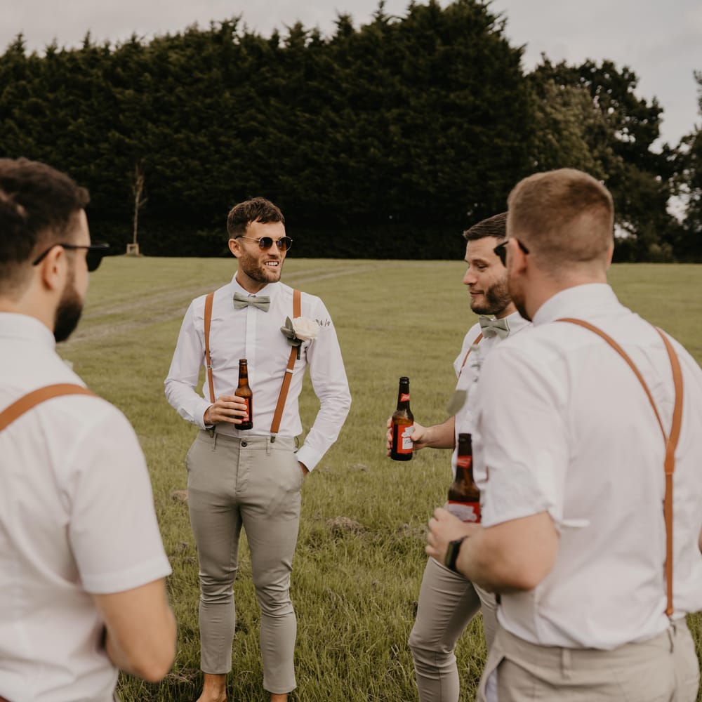 Groom & his groomsmen stand outdoors as they drink and groomsmen wear shirts with brown braces | Mark Bamforth Photography