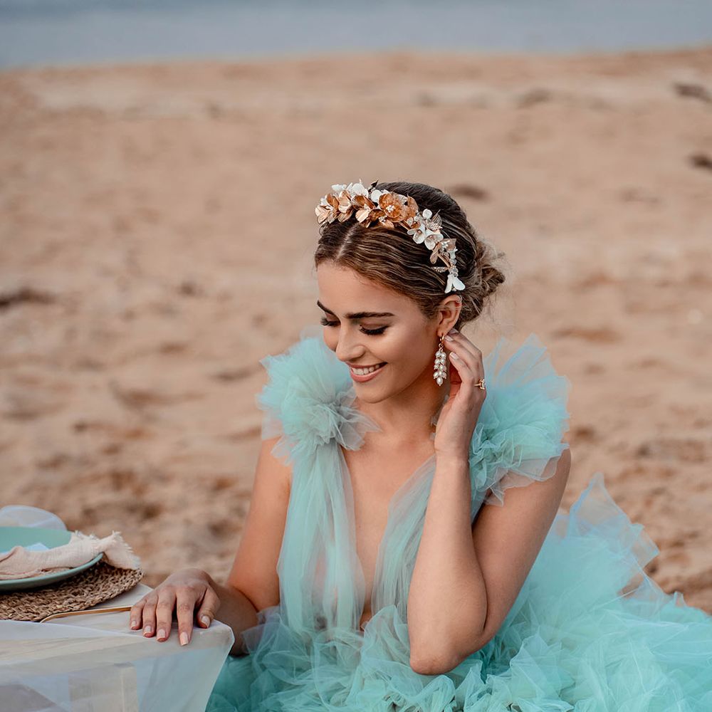Bride in a mint green wedding dress, gold headband and dangly earrings sitting at her relaxed beach reception 