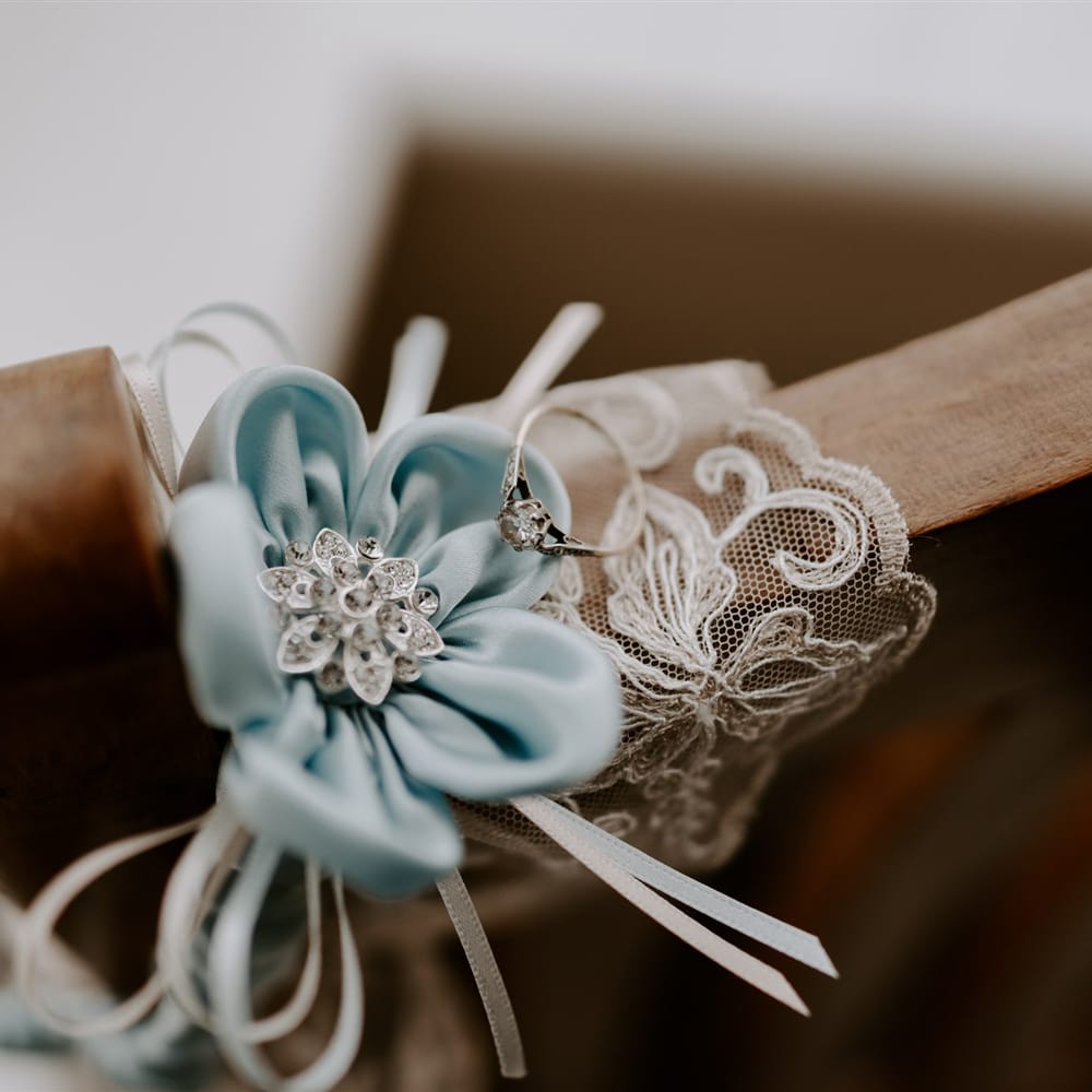 Blue silk flower with silver centre and diamond wedding ring resting on wooden chair before DIY garden wedding in Bedfordshire
