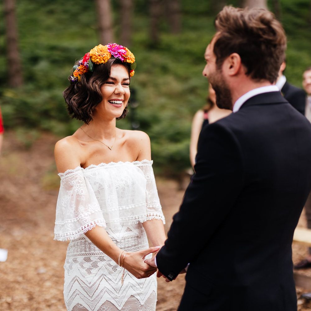 Bride in strapless Grace Loves Lace wedding dress and colourful flower crown laughs during wedding ceremony whilst holding hands with groom in black suit at outdoor wedding ceremony
