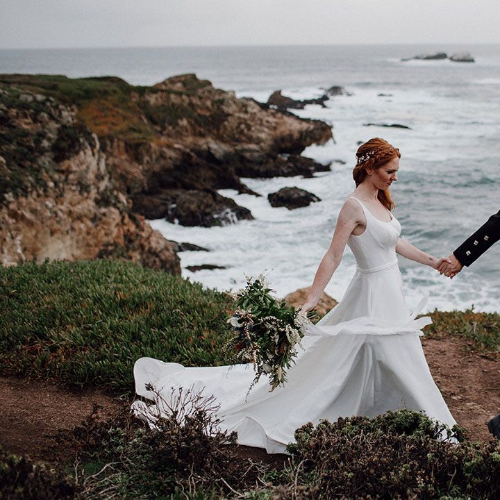 Groom in a tartan kilt holding hands with his bride in a floaty wedding dress as they walk along the cliff edges at Big Sur