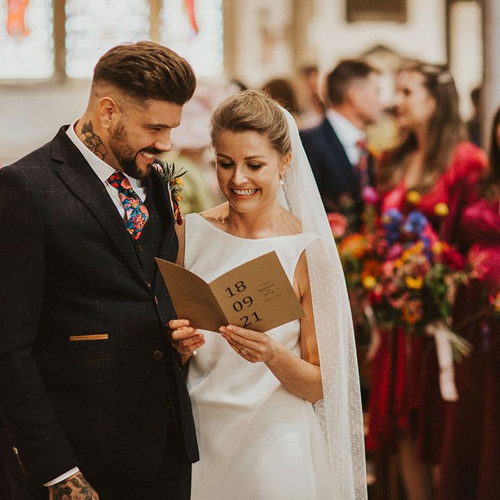 Bride in a slip wedding dress and cathedral length veil and groom in a three piece wool suit holding a craft card order of service at their church wedding ceremony 