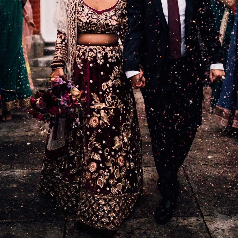 Bride in burgundy Indian saree wedding dress walks out of wedding with groom in dark suit and burgundy tie as guests throw confetti 