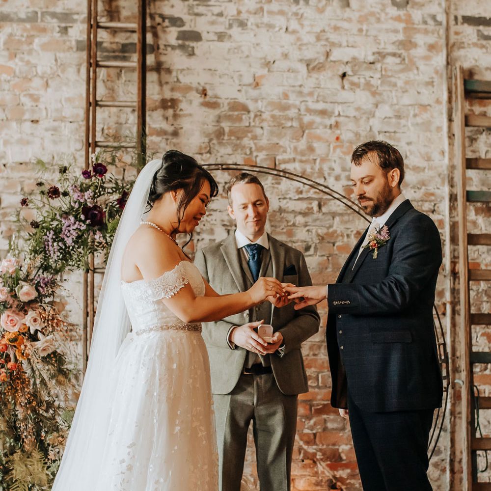Bride in floral lace wedding dress and veil puts ring on hand of groom in blue three piece suit and brown shoes in front of floral moongate during wedding ceremony