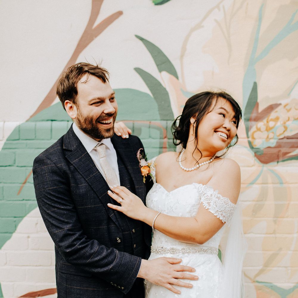 Groom in three piece check suit with light pink tie stands with bride in sweetheart neckline lace wedding dress and pearl necklace