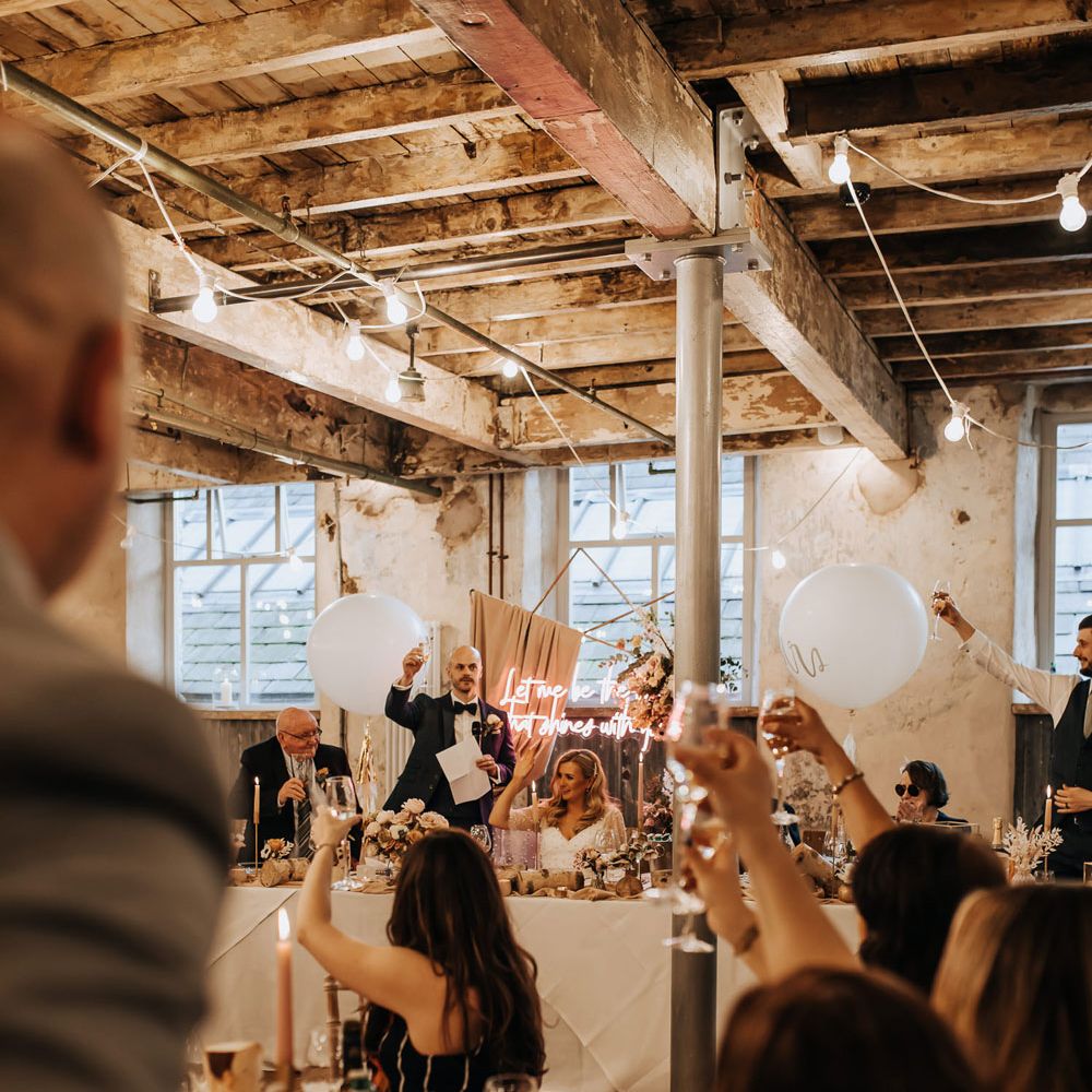 Guests raise a glass during wedding reception in industrial mill wedding venue with rustic styling and neon wedding sign