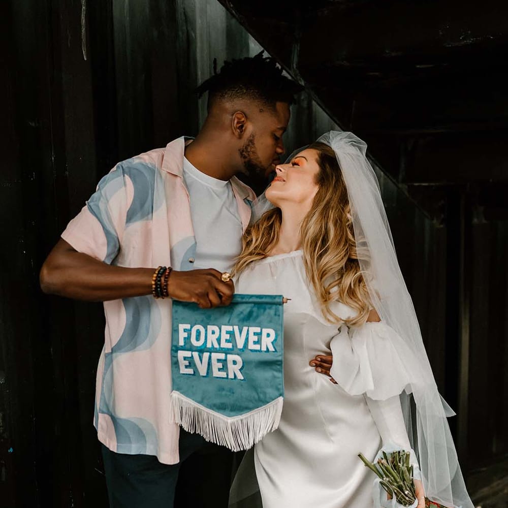 Groom in a pastel patterned shirt holding a Forever Ever teal suede fabric wedding banner 