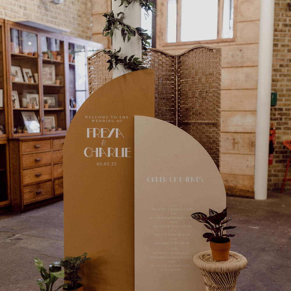 Wedding welcome sign and oder of events notice board in deep mustard and cream with potted plants and wicker details