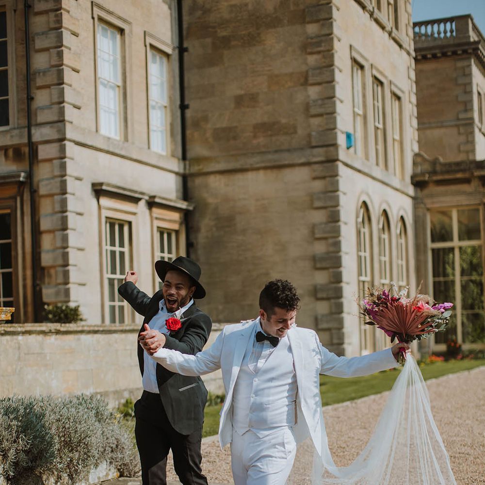 Groom in a two piece black suit, fedora hat and boots holding hands with his partner in a white three piece groom suit, white boots and cape 