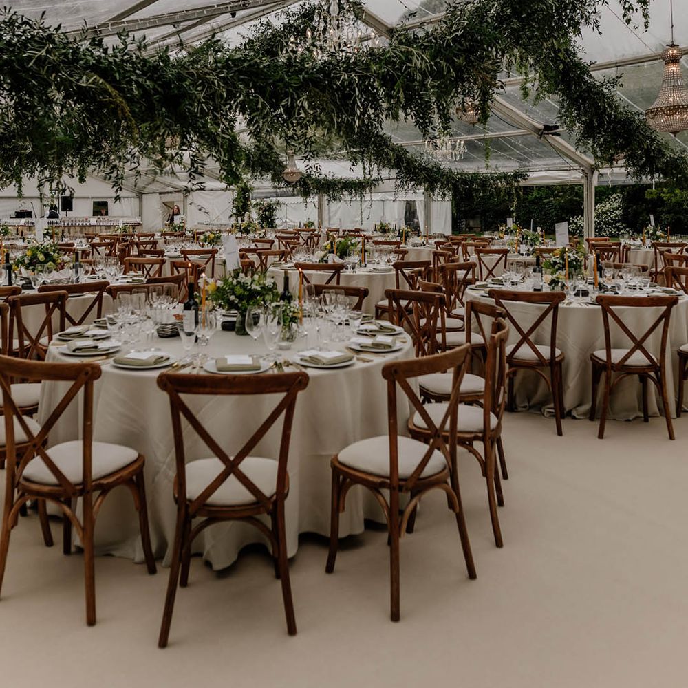 Clear glass marquee wedding reception with wooden furniture and hanging greenery garlands 