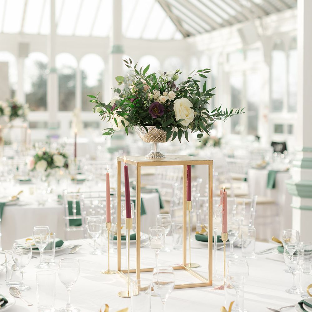Floral bouquet sits in vase upon gold stand surrounded by candles in burgundy and pale pink 