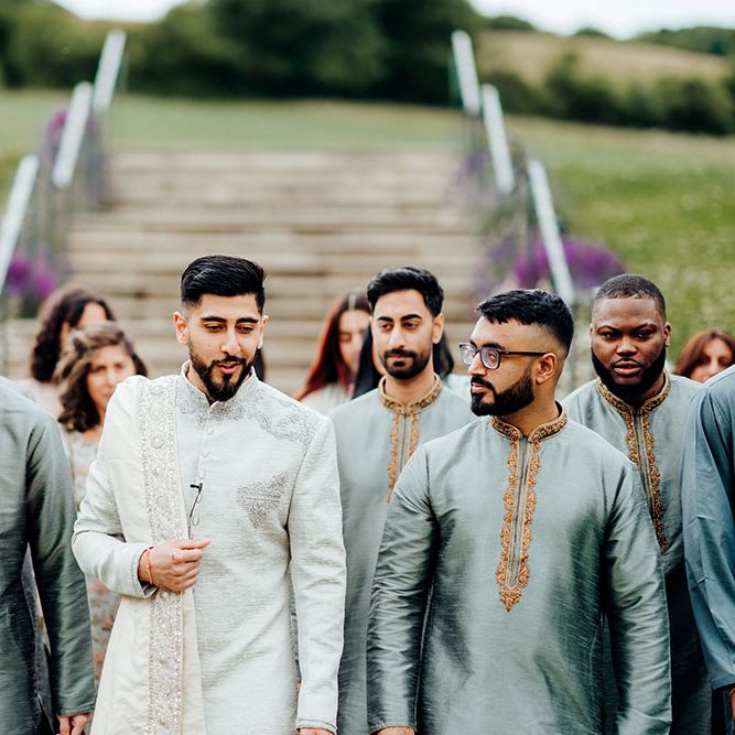 Groom wearing traditional light grey sherwani walks with his groomsmen who wear deep grey sherwanis with gold embellishment 